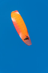 A single paraglider with a vibrant orange and red canopy is seen against a deep, clear blue sky during a sunny day flight.