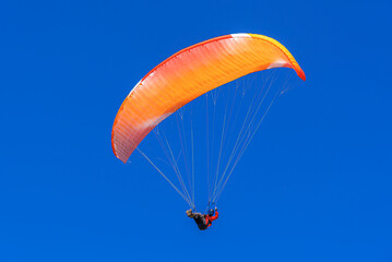 A single paraglider with a vibrant orange and red canopy is seen against a deep, clear blue sky during a sunny day flight.