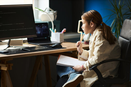 Caucasian preteen girl sitting at desk studying computer code on monitor, holding notebook and pen, appearing focused while learning programming in modern home office environment