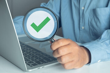 Quality Control (QA) and audit concept. Businessman holding magnifying glass over green check mark icon on laptop. Inspection passed, verification, and ISO certification.