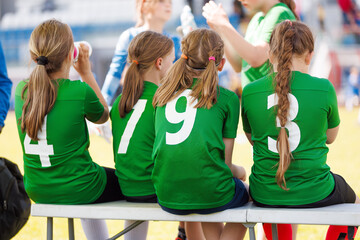 Girls in Green Jerseys Sitting on Bench. Girls Youth Soccer Team Taking a Break. Young Football Players in Green Jerseys Resting on Sideline Bench during Match © matimix