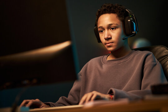 Portrait of Black teenage boy wearing headset sitting at desk, using computer monitor playing video game focused on screen hands on keyboard and mouse in gaming environment