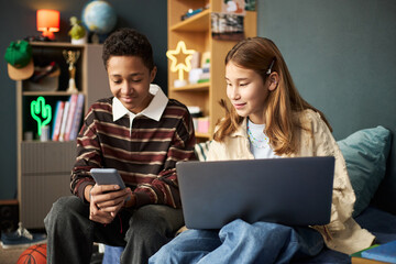 Teenage Black boy holding smartphone sitting next to teenage Caucasian girl using laptop, both smiling and interacting in modern bedroom with educational and personal items visible