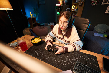 Teenage Caucasian girl wearing glasses sitting at desk playing video game with controller,...