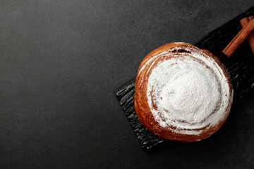 Round, delicious bun with cinnamon and powdered sugar on a black wooden board on a gray background