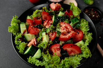 Vegetable salad of tomatoes, cucumbers, onions, lettuce dressed with aromatic dressing in a black plate on a gray background