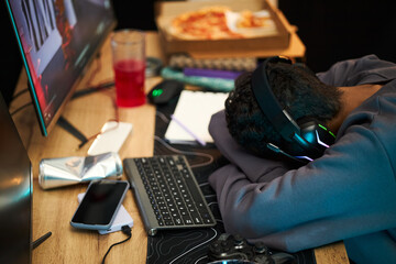 Young adult Black man wearing headphones sleeping at desk with head resting on arms near computer...