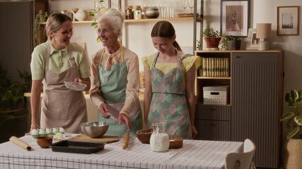 Happy family of senior grandmother, adult daughter and young granddaughter in aprons laughing while baking homemade cookies in kitchen spending time together on weekend, slow motion