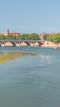Garonne River and Pont Neuf timelapse in downtown Toulouse, France. Renaissance arch bridge reflects in the water under a blue sky with Basilica of Our Lady of the Daurade. Waterfront with green trees