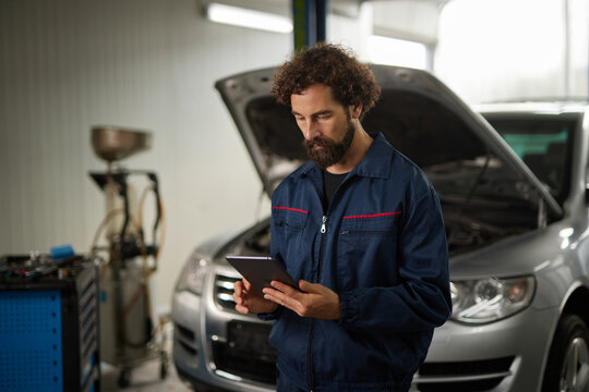 A mechanic examines vehicle data on a tablet while standing in a car repair shop. The car hood is open, and tools are visible in the background.