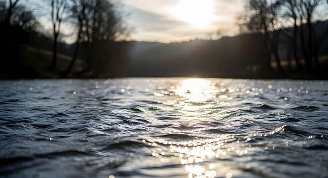 Sunlight glistening on ripples of a tranquil river at dusk and surrounded by silhouetted trees and a gentle slope under a soft sky
