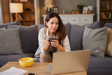 A woman sits comfortably on her couch, holding a cup of coffee and looking at her laptop screen. A bowl of snacks rests on the table beside her.