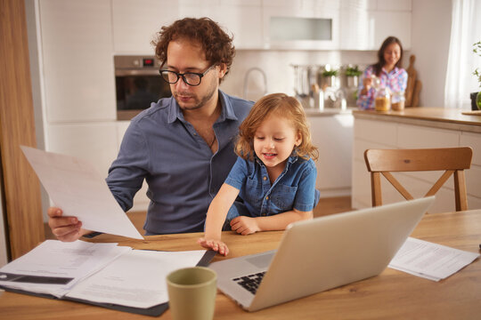 A father reviews paperwork at the kitchen table while his daughter happily engages with him. Their mother prepares breakfast in the background, creating a warm family atmosphere.
