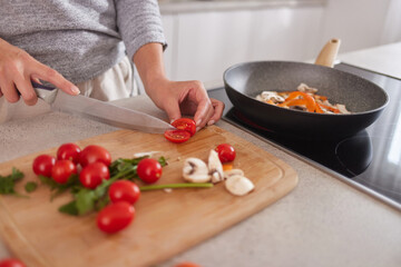A person is chopping cherry tomatoes on a wooden cutting board in a bright kitchen. Fresh mushrooms and bell peppers are cooking in a pan nearby, creating a vibrant meal.