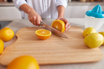 A person is cutting an orange on a wooden board in a sunny kitchen. Fresh lemons and additional oranges surround the cutting area, creating a vibrant scene of food prep.