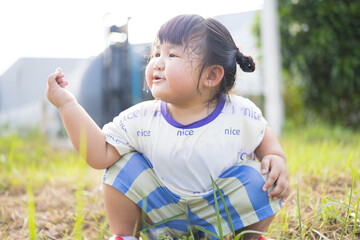 A cute little Asian girl is sitting on the grass and making a funny face,Cheerful Asian girl with cute bows sitting cross-legged on green grass, enjoying a sunny day outdoors.