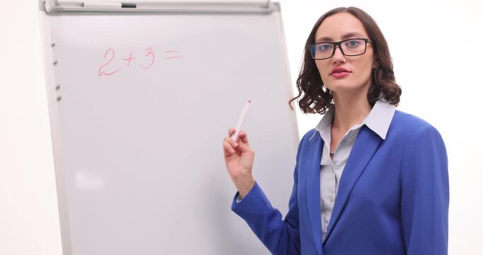 A teacher in a blue jacket explains basic math concepts, writing on a whiteboard in a well-lit classroom. She guides students through problem-solving with enthusiasm.