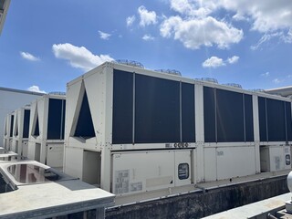 A row of large, modular commercial air-cooled chillers and HVAC units sits on a rooftop under a bright, partly cloudy blue sky. These systems are essential for industrial climate control and building 