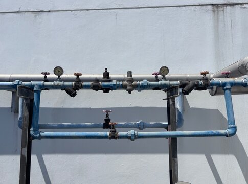 Industrial pipework system on an exterior building wall, featuring a backflow preventer assembly, gate valves, and pressure gauges on galvanized and blue-painted water pipes under bright sunlight.