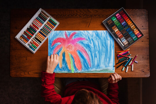 Overhead view of a young boy sitting at a table with assorted multi coloured chalks and crayons drawing a picture