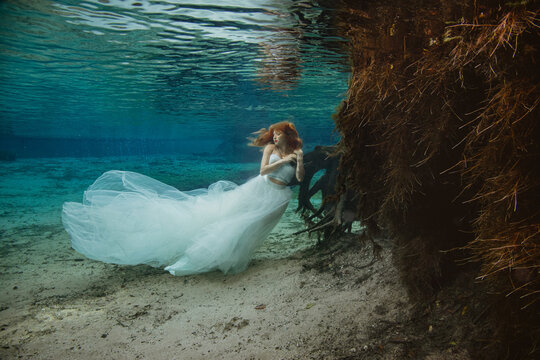 Beautiful redheaded woman in a strapless beaded top and long skirt sitting underwater on a riverbed holding the roots of a tree with her eyes closed, Florida, USA