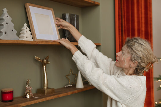 Close-up side view of a Woman arranging an empty picture frame on a shelf amongst assorted festive Christmas decorations