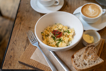 Close-up of two cappuccinos and a bowl of scrambled eggs, toast and butter on a wooden table