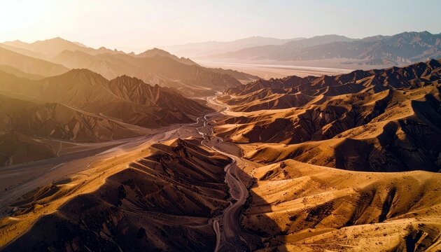 Aerial view of arid, sun-drenched desert mountains with dramatic shadows, a winding road, and a dry riverbed stretching into the distance.