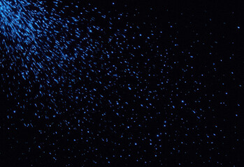 Abstract dark background with blue particles scattered across space, creating a shimmering starry trail and cosmic dust texture for design projects, digital art and presentations