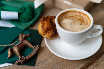 Close-up of a cup of coffee with a sweet pastry on wooden table with a rocking horse ornament, wrapped Christmas gift and stationery
