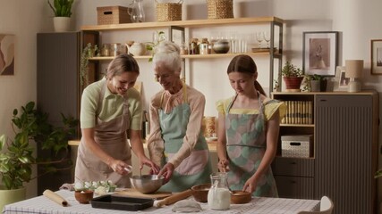 Adult woman in apron cracking egg into mixing bowl making batter for crepes with elderly mother and young daughter while sharing warm moment in cozy kitchen, slow motion - Powered by Adobe