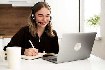 woman operator with headset writing notes in notebook working remotely in the kitchen