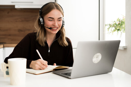 woman operator with headset writing notes in notebook working remotely in the kitchen