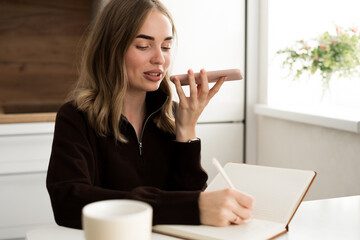 young student freelancer working and recording voice message on smart phone, writing notes in notepad