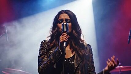Talented female singer performing on a stage during a live music concert, holding a retro microphone with smoke and bright lights in the background while her band is playing instruments - Powered by Adobe
