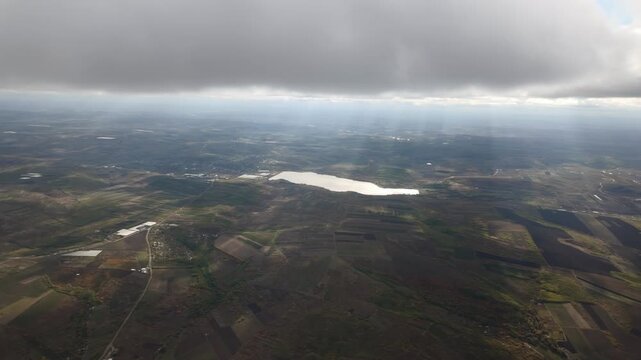 Aerial view, flying over green fields at level of cumulus clouds with sun rays, fly into gray cloud. View from clouds on green meadows with lake shining in sunshine. Natural cloudscape with sunbeams