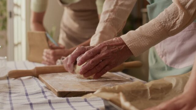 Close up of grandmothers hands kneading dough on wooden board teaching adult daughter and granddaughter homemade puff pastry recipe while cooking together in kitchen