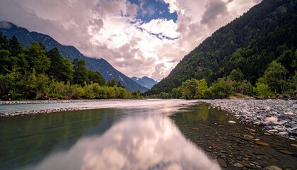 A wide, slow-moving river winds through a valley, flanked by steep, forested mountains and a rocky shoreline, with dramatic clouds in the sky.