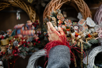 Hands holding a decorative Christmas wreath with pine cones, ribbons, and festive ornaments in a holiday shop.