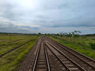 Fototapeta premium Two sets of parallel railroad tracks converging in the distance, surrounded by green trees, grass, and dry earth, with residential buildings visible in the background under a moody cloudy sky.