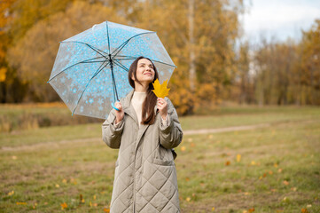 Smiling woman with umbrella holding yellow maple leaf in autumn park