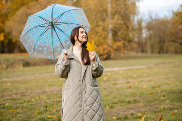 Smiling woman with umbrella holding yellow maple leaf in autumn park