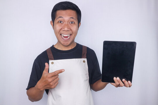 Smiling young Asian man barista barman employee wear apron hold digital tablet showing empty screen