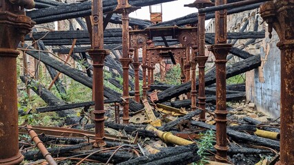 Burned down industrial factory building, charred collapsed wooden roof beams. Rusting burnt twisted metal debris. Derelict warehouse arson aftermath. Columns ruins, support pillars after fire damage.