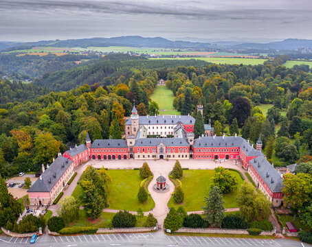 Aerial view of Sychrov Castle's red and white facade amidst autumn's vibrant forest, a historic gem under a cloudy sky, Sychrov, Liberecky kraj, Czechia.