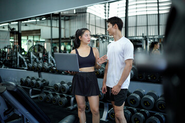 Asian man and woman exercise training in fitness gym. Young female trainer using laptop for schedule exercise training to fitness male. Workout training in fitness gym.