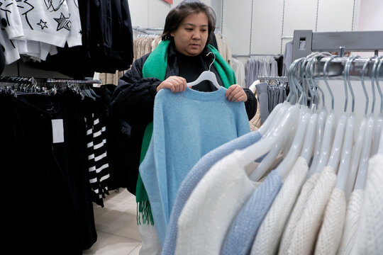 A woman is browsing sweaters in a retail clothing store, holding a light blue sweater and examining it. Racks of various sweaters and garments are visible around her.