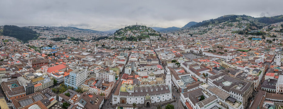 Aerial panoramic view of a sprawling cityscape unfolds beneath a cloudy sky, revealing the Metropolitan Cathedral and the distant Panecillo, Quito, Ecuador.