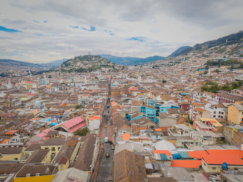 Aerial view of terracotta rooftops cascade down towards narrow streets, shadowed by the distant Panecillo hill topped with its iconic statue, Quito, Ecuador.