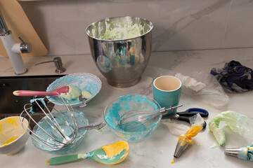 A pastry chef's workspace is filled with colorful bowls of meringue and whipped cream, along with mixing tools. The countertop is cluttered with baking supplies and remnants from the sweet creations.
